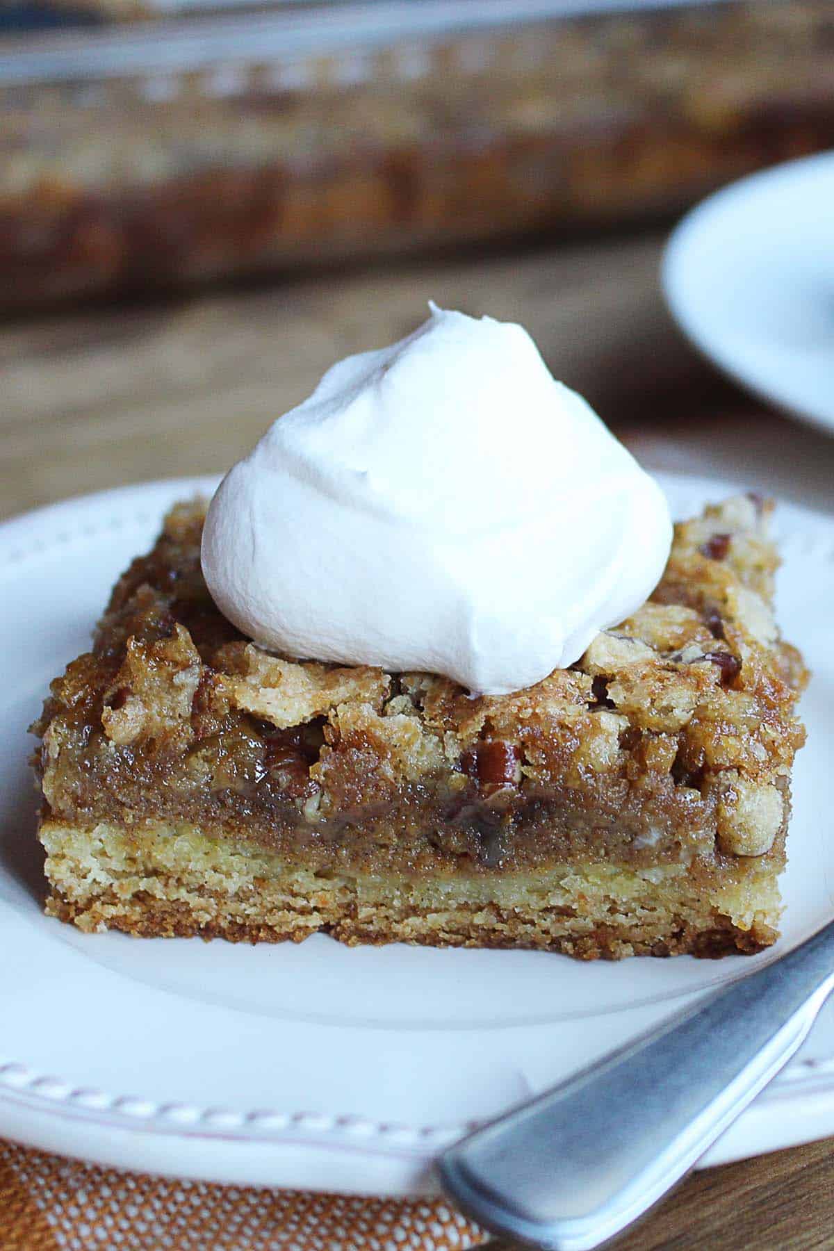 pumpkin-gooey-butter-crunch-cake-featured-1 Pumpkin gooey butter crunch cake has a cake layer, pumpkin filling, and pecan filled topping. The slice is on a white plate with a dollop of whipped cream on top.