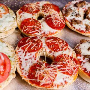 Tray of hot, melted freezer-friendly pizza bagels with pepperoni, melted mozzarella cheese, and crispy edges. In the background are margherita and sausage pizza bagels.