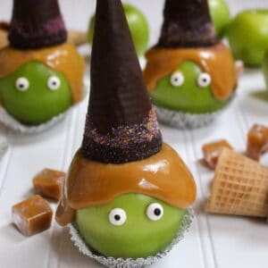 Five witch caramel apples topped with chocolate sugar cone witch hats and Halloween sprinkles sitting in cupcake liners