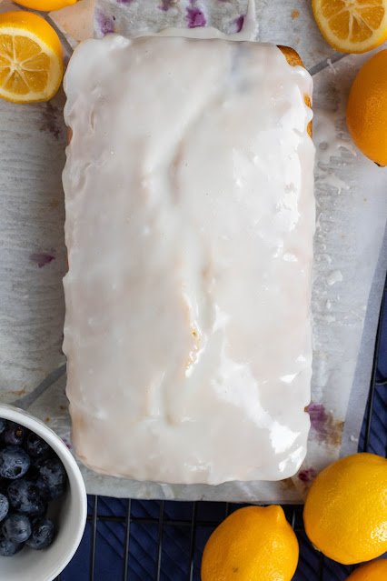 Unsliced loaf showing the lemon icing covering the Whole Lemon Blueberry Cake. Fresh blueberries and lemons are in the background.