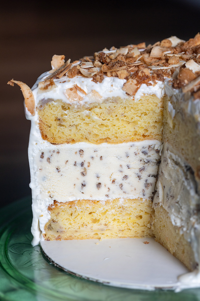 A whole homemade banana coconut ice cream cake frosted with smooth whipped cream and decorated with toasted coconut flakes and chopped macadamia nuts.