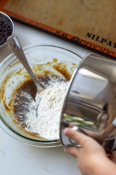 Little girl pours bowl of dry ingredients into bowl of wet ingredients for chocolate chip ice cream sandwich cookies