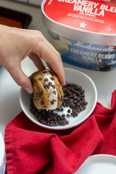 A hand holding a chocolate chip ice cream sandwich cookie being dipped into mini chocolate chips.