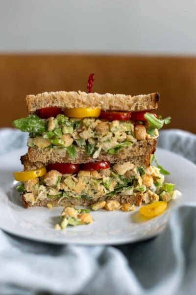 Two halves of a mashed chickpea salad sandwich stacked on a plate showing the textured chickpea filling, sliced tomatoes, and leafy green arugula.