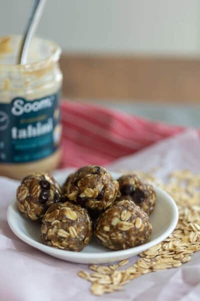 A small white bowl filled with six rounded nut-free energy bites, made with oats, tahini, and mini chocolate chips. The bowl sits on parchment paper next to a jar of tahini and scattered dry oats.