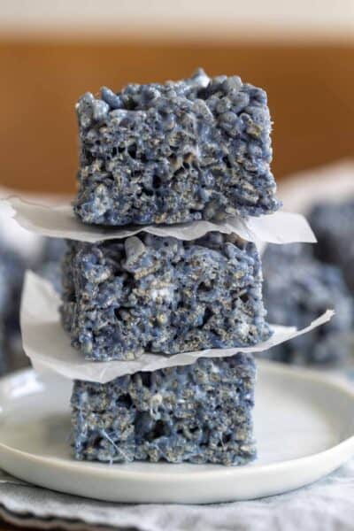Three thick, square-cut blue Rice Krispies treats stacked high on small squares of parchment paper on a white ceramic plate.