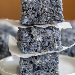 Three thick, square-cut blue Rice Krispies treats stacked high on small squares of parchment paper on a white ceramic plate.