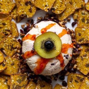 Eyeball cheese ball using a kiwi slice and blueberry to create the iris and pupil. Red pepper jelly is drizzled over the eyeball to make the bloody appearance. This is surrounded by pumpkin crackers.