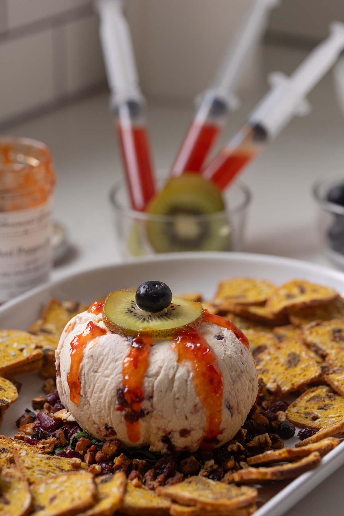 Eyeball cheese ball using a kiwi slice and blueberry to create the iris and pupil. Red pepper jelly is drizzled over the eyeball to make the bloody appearance. This is surrounded by pumpkin crackers.