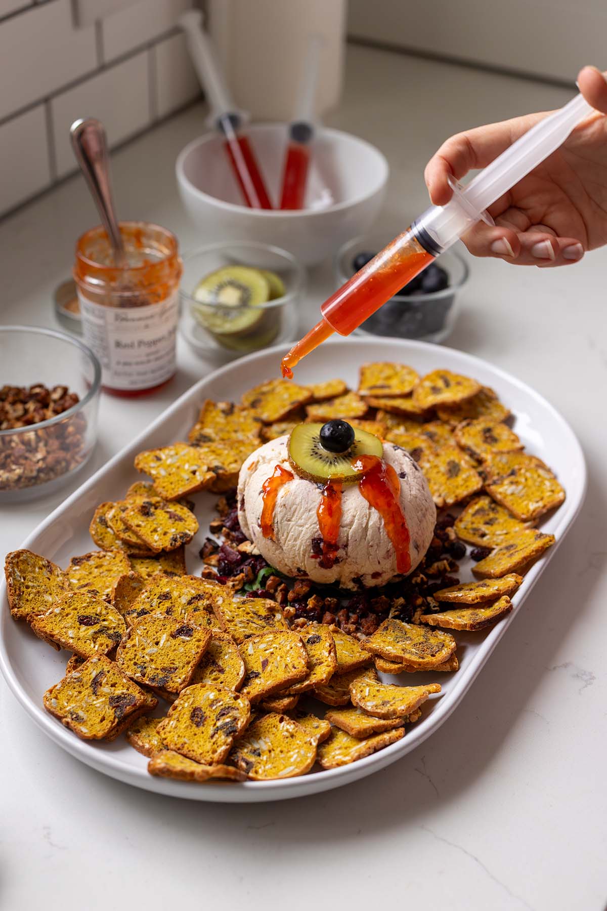Eyeball Cheese Ball Syringe Technique Eyeball cheese ball using a kiwi slice and blueberry to create the iris and pupil. Red pepper jelly is drizzled over the eyeball using a food grade syringe to make the bloody appearance. This is surrounded by pumpkin crackers.